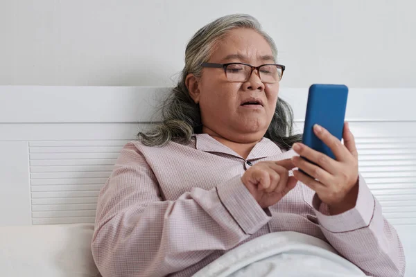ショックを受けた先輩女性がベッドに座ってスマートフォンでニュースを読む — ストック写真