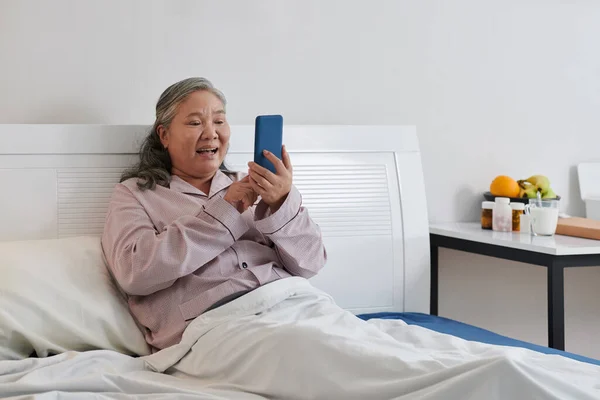 快乐的老年妇女坐在床上 给孩子们打视频电话 — 图库照片