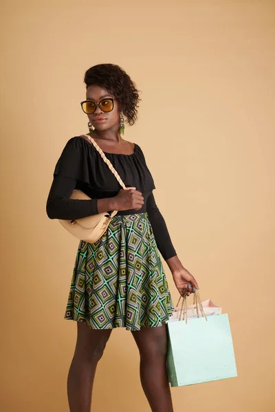スタイリッシュな自信に満ちた黒の若い女性でサングラスを運ぶショッピングバッグと離れて見る — ストック写真