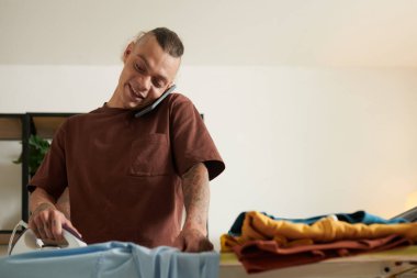 Neşeli genç adam elbiselerini ütülüyor ve telefonda arkadaşlarıyla haberleri tartışıyor.