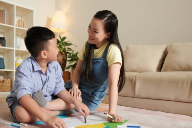 Gülümseyen Asyalı anne ve oğlu yerde oturuyor, resim çiziyor ve boyuyor.