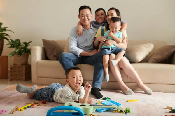 彼の両親と兄弟がソファに座って抱き合っているときに小さな男の子が床に横たわって舌を突き出す — ストック写真