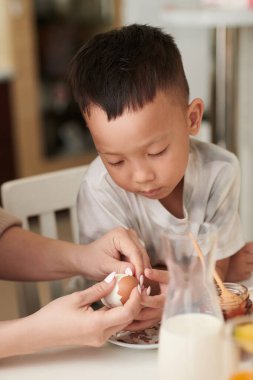 Meraklı küçük çocuk haşlanmış yumurta soyan annesinin ellerine bakıyor.