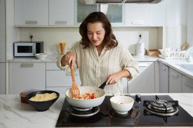 Gülümseyen kadın akşam yemeği için spagetti sosu pişirirken kızarmış sebze kokusundan zevk alıyor.