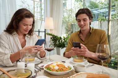 Romantik bir akşam yemeğinde birbirleriyle konuşmak yerine arkadaşlarına mesaj atan ya da sosyal medyayı kontrol eden genç bir çift.