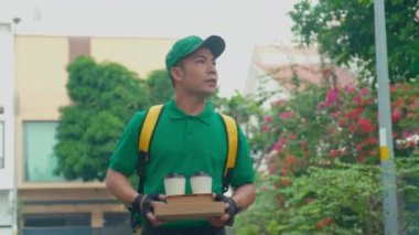 Yeşil üniformalı, şapkalı, sarı çantalı, kahve kolili bir teslimatçı. Çiçek açan arka plandaki müşteri adreslerini arıyor.