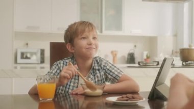 Küçük çocuk kahvaltısını mısır gevreği, meyve suyu ve kurabiyeyle yaparken annesiyle konuşuyor ve dijital tablette çizgi film seyrediyor.