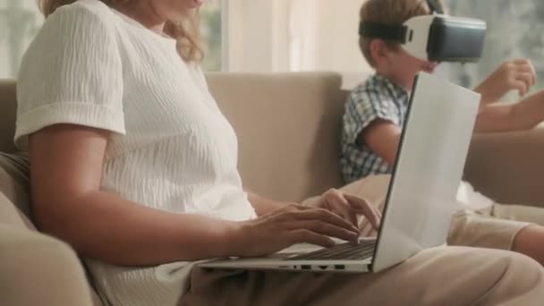 息子を使用してVr メガネゲームをプレイしながら 母親はリビングルームのソファの上に座ってラップトップで作業します — ストック動画