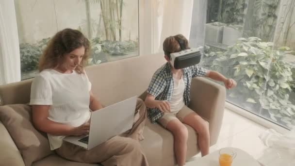 儿子用Vr眼镜玩游戏的放大镜头和妈妈坐在客厅沙发上用笔记本电脑工作的镜头 — 图库视频影像