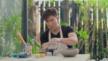 Asyalı bir adam atölyede çömlek çarkına kil çömlek oyuyor.