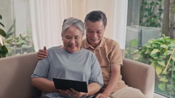 在客厅的沙发上 坐在一块数字平板电脑上的一对老夫妇一边微笑一边上网的照片 — 图库视频影像