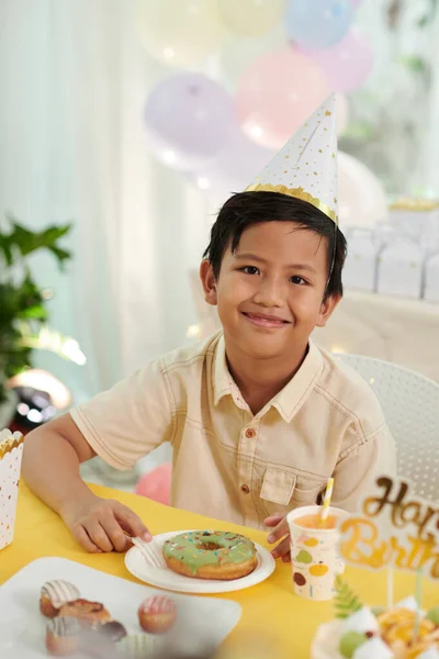ドーナツを食べるときにパーティの帽子をかぶった笑顔の男の子の肖像画 — ストック写真