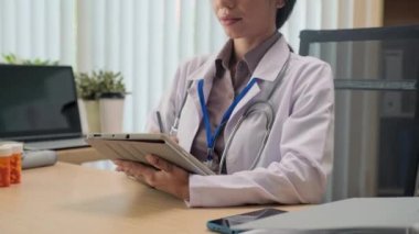 Kadın doktorun dijital tablet üzerinde çalışırken ve klinikteki iş yerinde gülümserken kameraya poz verirken görüntüsünü kaldır