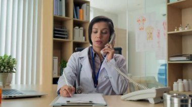Kadın doktorun telefon görüşmelerine cevap verirken ve tıbbi kartıyla not alırken orta boy bir fotoğrafı.