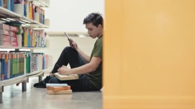 Erkek öğrencinin kütüphane zemininde oturup dijital tablet üzerinde notlar alıp gelecek projeye hazırlandığı uzun bir çekim.
