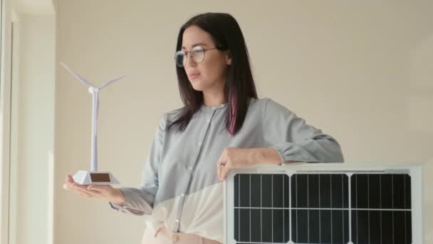 一个快乐的年轻女子的画像 她拿着风力涡轮机塑料模型 靠在太阳能电池板上 看着室内相机 — 图库视频影像