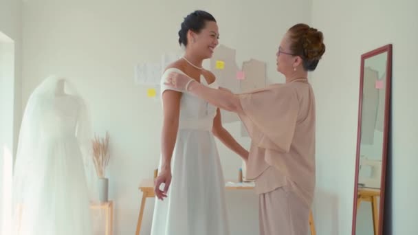 中长镜头的母亲看着幸福的女儿在工作室里穿上白色婚纱 — 图库视频影像