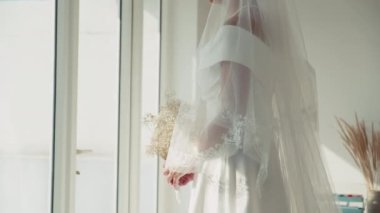 Beyaz gelinlik ve duvak giymiş gelinin, pencerenin önünde bir buket çiçekle salonda dikilirken çekilmiş resmi.