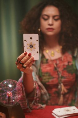 Tarot kartları okuyucusu elinde büyük bir arcana bakıyor, seçici bir odaklanma.