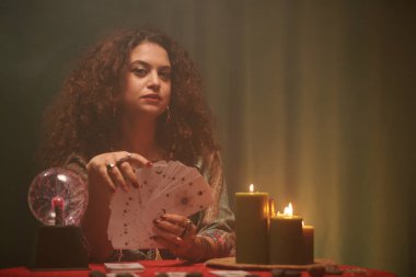 Tarot kartlı falcının portresi yanan mumlar ve kristal küreyle masada oturuyor.