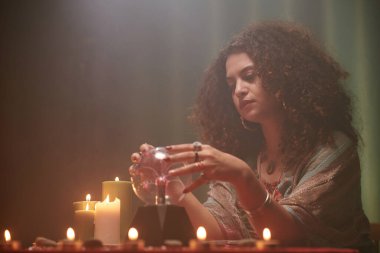 Düşünceli büyücü, yanan mumlarla masada otururken kristal küreye bakıyor.