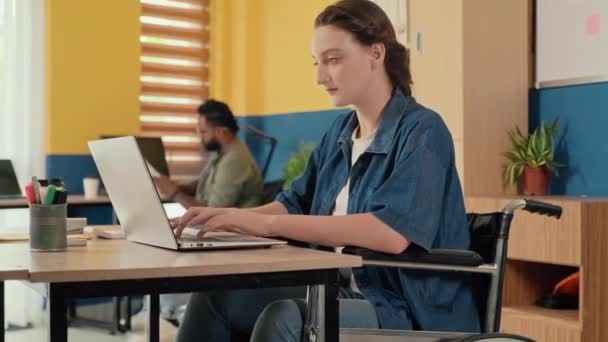 坐在轮椅上的年轻女性在笔记本电脑上工作和在办公室看着相机的画像 — 图库视频影像