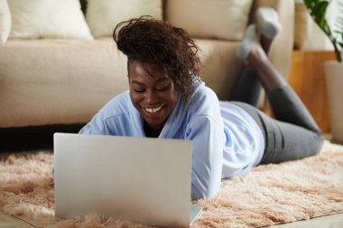 Genç ve mutlu bir kadın yerde yatar ve çalışır ya da dizüstü bilgisayardan yeni bir program izler.