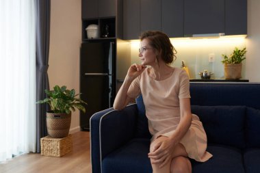 Gülümseyen olgun kadın evdeki kanepede oturuyor ve başka tarafa bakıyor.