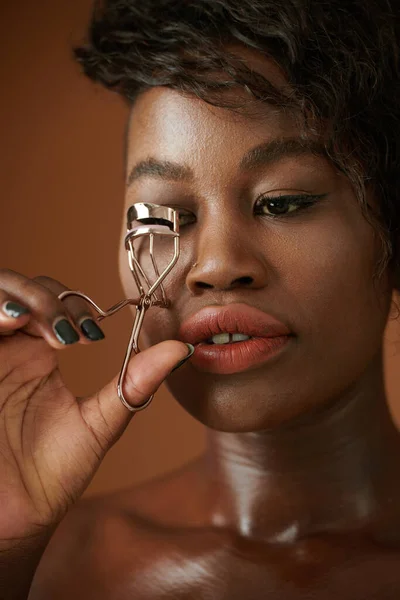 Young Black woman curling lashes with tool when doing make-up for event or party