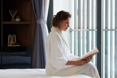 Olgun bir kadın rahat bir bornozla yatakta oturuyor ve büyüleyici bir roman okuyor.