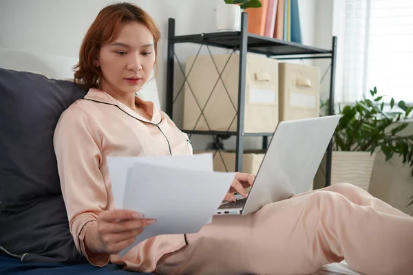 Young transgender businesswoman sitting on bed in pajamas and working with documents