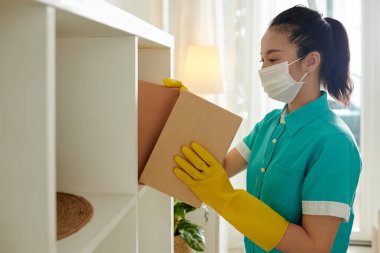 Tıbbi maskeli genç bayan temizlikçi tozları silmek için raflardan kitap alıyor.