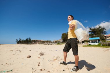 Kumsalda elinde çöp torbasıyla duran genç adam.