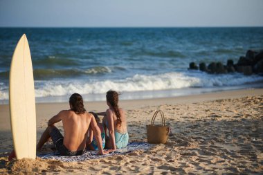 Suftboard 'lu bir aile kumsalda oturup deniz dalgalarını seyrediyor.