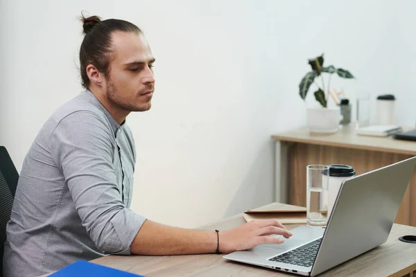 Serious young entrepreneur with man bun checking e-mails on laptop