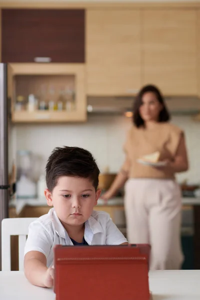 かわいい男の子は 母親がバックグラウンドで朝食を調理するときにタブレットコンピュータで漫画を見ています — ストック写真