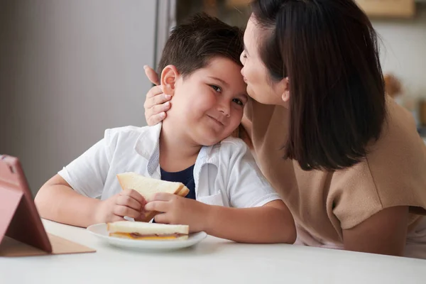 Mother kissing smiling son on forehead when he is eating sandwiches at kitchen counter