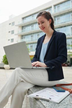 Neşeli iş kadını dışarıda oturuyor ve laptopla çalışıyor, iş arkadaşlarının mesajlarına ve e-postalarına cevap veriyor.