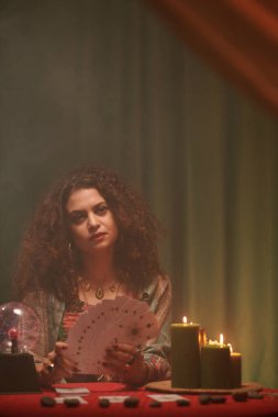 Masada yanan mumlarla otururken elinde tarot kartları tutan ciddi bir falcının portresi.