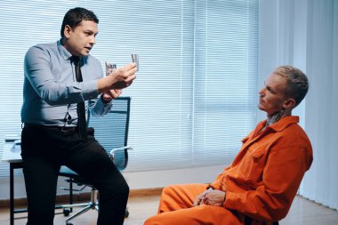 Dedektif mahkumu sorguluyor, ona olay yerinden fotoğraflar gösteriyor.