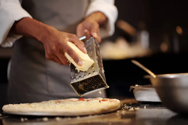 Hands of pizza maker shredding head of mozzarella cheese over small pizza