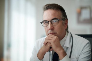 Ofisinde masasında oturan, işçi kıyafetli ve gözlüklü dalgın bir pratisyen doktorun portresi.