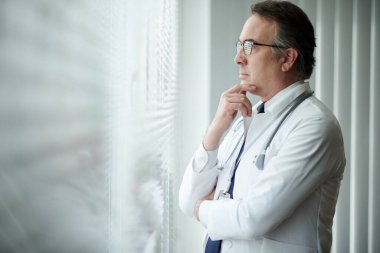 Pensive head of medical clinic rubbing chin and looking through office window clipart