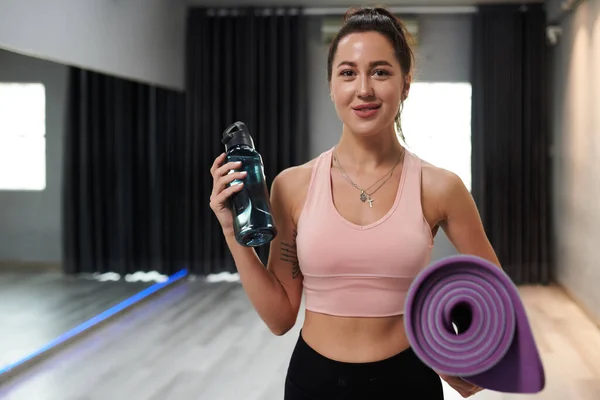 Portrait of fit cheerful fitness coach with bottle of water and yoga mat