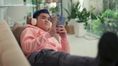 Pembe kapüşonlu mavi saçlı bir adamın akıllı telefonuyla kanepede dinlenip kulaklıkla müzik dinlerken orta boy fotoğrafı.