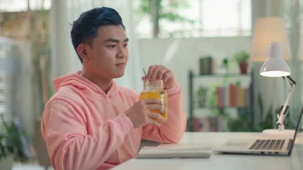 ピンクのフーディーの青い髪の若い男は ラップトップとテーブルに座ってレモネードを飲んで 自宅で作業し ノートを書いています — ストック動画
