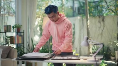 Orta boy, pembe kapüşonlu mavi saçlı fotoğrafçı yarışmaya hazırlanırken çerçeveye resim koyar, stüdyoda masa başında çalışır.