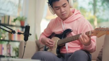 Kulaklıklı, mavi saçlı genç Asyalı adamın gitarını dizüstü bilgisayarla çalarken görüntüsünü kaldır.
