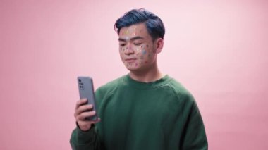 Pembe arka planda durup kameraya bakarken fotoğrafta mavi saçlı ve yüzünde çıkartmalar olan bir fotoğrafçı akıllı telefondan selfie çekiyor.