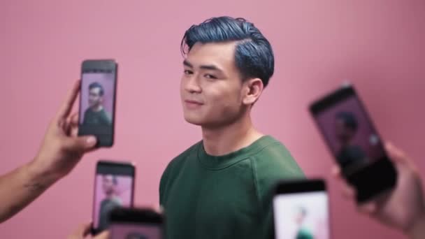 青い髪の陽気なアジアのブロガーのミディアムショットは ピンクの壁の背景に立っている間 スマートフォンのカメラで彼のファンの多くによって撮影されている楽しんでいます — ストック動画